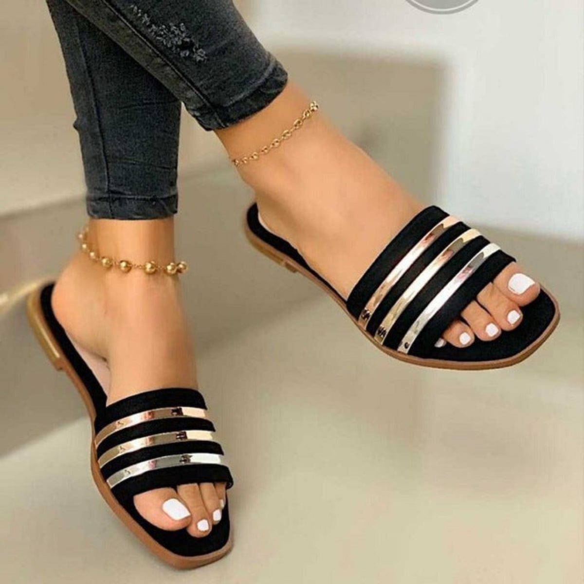 2black women’s flat slide sandals featuring multiple horizontal black straps accented with reflective metallic silver-tone stripes, an open-toe silhouette, slim flat sole, and easy slip-on design, shown worn on both feet from a close front angle to clearly display the striped upper and overall fit.