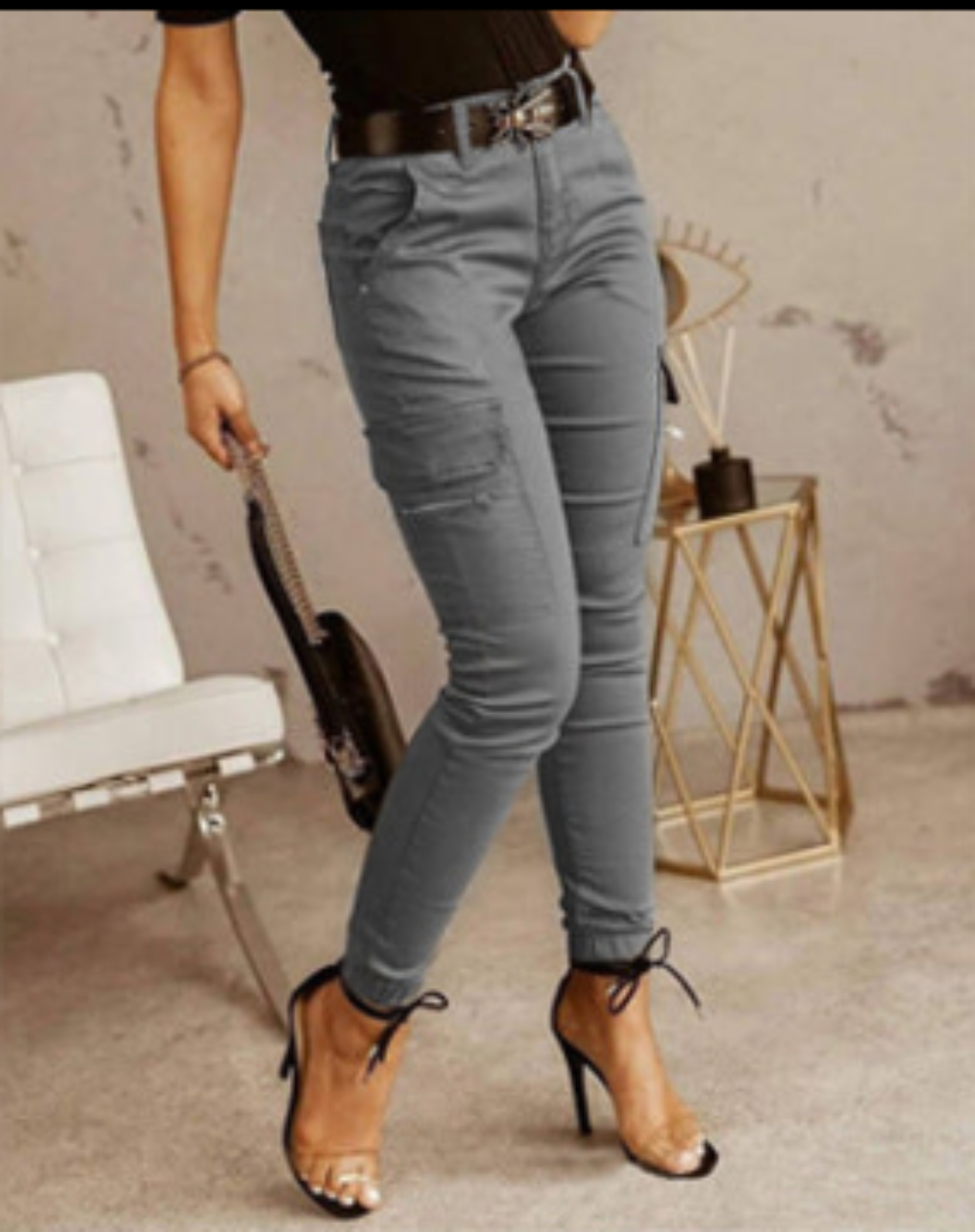 A person wearing slim-fit gray cargo-style jeans with a black belt, paired with a black top and tan heeled sandals, standing in a styled indoor setting with a white chair and gold side table.