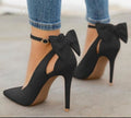 Black Arielle Bow Ankle-Strap Stiletto Pumps worn with cropped jeans, featuring large sculpted ankle bows, sleek pointed toes, slim stiletto heels, and an elegant all-black finish.