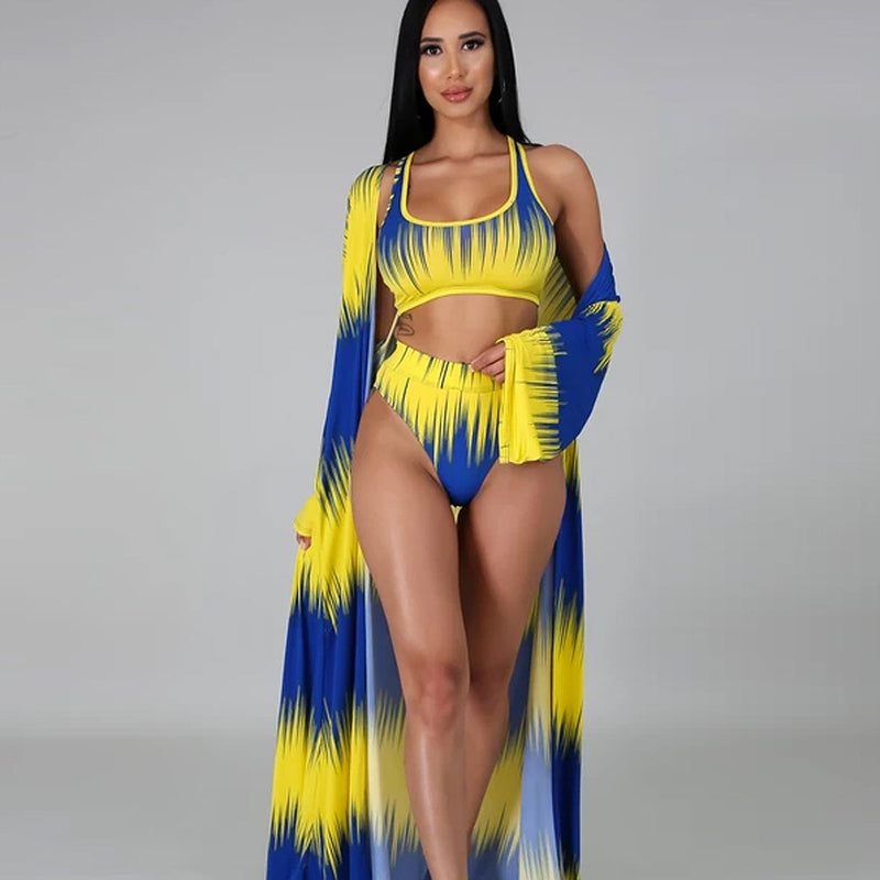 Azaria women's three-piece resort swimwear set shown on a seated model, featuring a sleeveless blue-and-yellow gradient-print cropped swim top, matching high-waisted bikini bottoms, and a long coordinating cover-up with dramatic yellow streaks transitioning into royal blue.