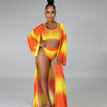 Full-body view of a model wearing the Azaria orange-and-yellow three-piece swimwear outfit, featuring a bright yellow cropped swim top with flame-like orange gradient detailing, matching high-waisted bikini bottoms, and a long flowing orange-and-yellow printed cover-up worn open.