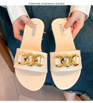 Beige women's flat slide sandals with a wide smooth strap, oversized polished gold-tone chain buckle embellishment, open-toe design, and light beige footbed, shown as a matching pair being held over blue denim.