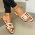 Beige women’s flat slide sandals featuring multiple horizontal beige straps with polished metallic stripe accents, an open-toe silhouette, slim flat sole, and easy slip-on design, shown worn on both feet from an angled front view to highlight the neutral color, metallic detailing, and streamlined summer style.