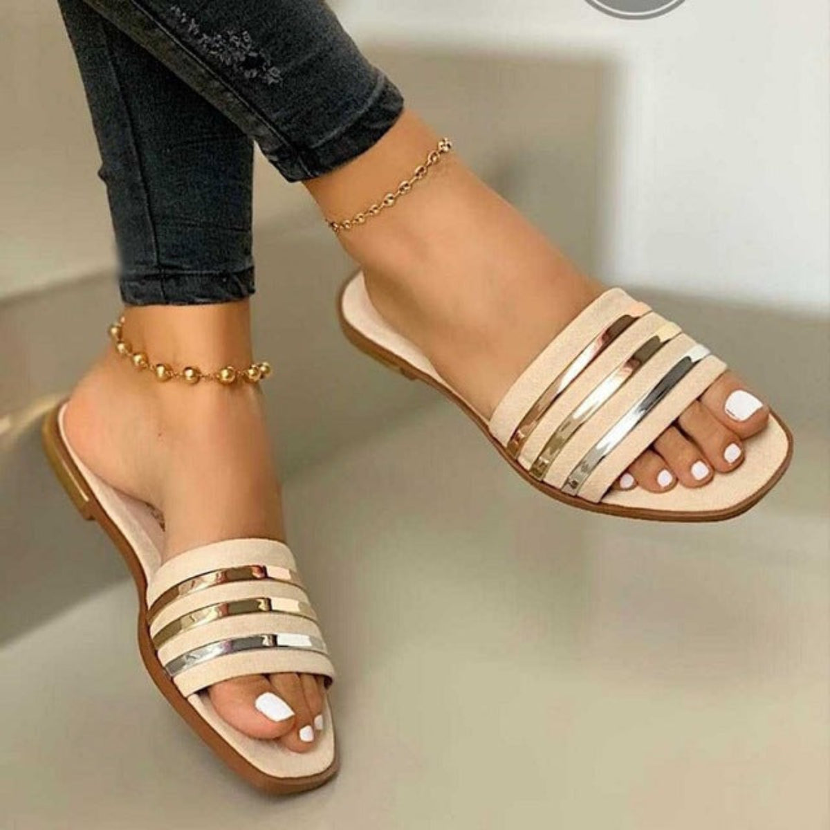 Beige women’s flat slide sandals featuring multiple horizontal beige straps with polished metallic stripe accents, an open-toe silhouette, slim flat sole, and easy slip-on design, shown worn on both feet from an angled front view to highlight the neutral color, metallic detailing, and streamlined summer style.