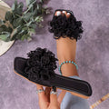Black women’s flat slide sandals featuring dramatic layered 3D flower embellishments across the upper strap, an open-toe silhouette, and low flat sole, shown from an angled product view highlighting the raised floral texture and slip-on construction.