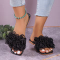 Black women’s 3D floral flat slide sandals shown worn from the front, featuring oversized dimensional black flower embellishments across the upper straps, open toes, and an easy flat slip-on design styled with cropped light-wash denim.