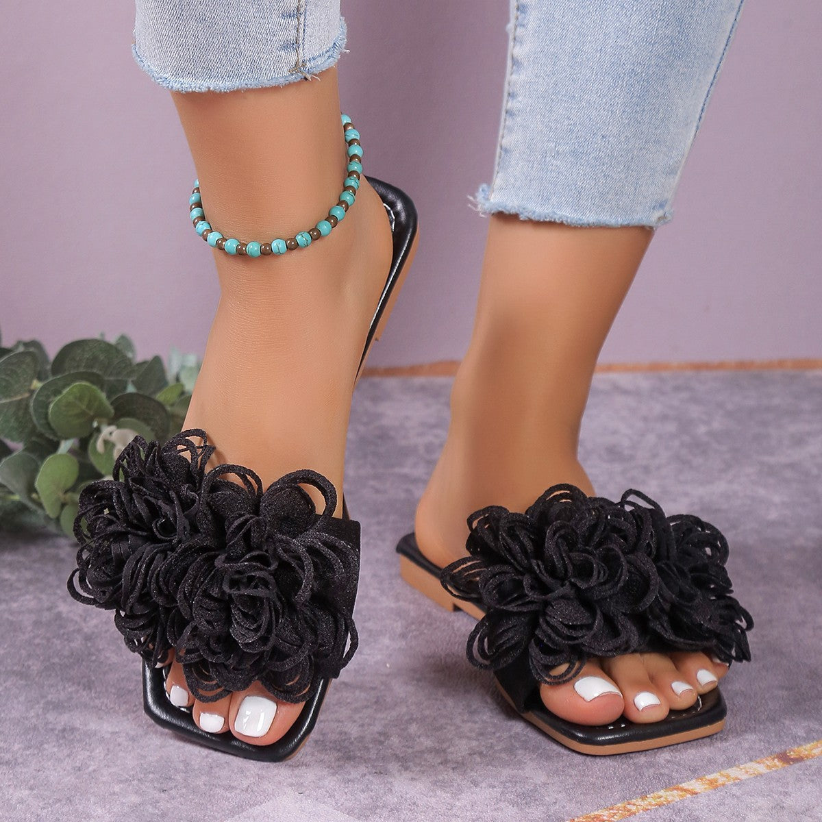 Black women’s 3D floral flat slide sandals shown worn from the front, featuring oversized dimensional black flower embellishments across the upper straps, open toes, and an easy flat slip-on design styled with cropped light-wash denim.