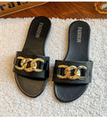 Black women's flat slide sandals featuring a wide black strap, oversized polished gold-tone chain buckle detail, open-toe silhouette, and matching black footbed, displayed as a pair on a soft cream textured rug.