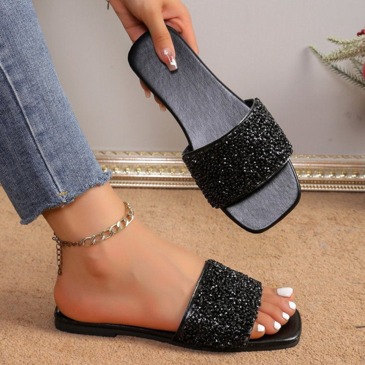 Black women’s flat slide sandals featuring a wide upper strap covered in sparkling black rhinestone embellishments, an open-toe silhouette, slim black flat sole, and easy slip-on design, shown worn on one foot while the matching sandal is held beside it to highlight the glittering texture and overall shape.
