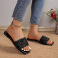 Black women’s rhinestone embellished flat slide sandals shown worn from an angled side view, featuring a wide glittering black upper strap, open-toe silhouette, matching black footbed, and slim flat sole styled with cropped denim for an elevated casual look.