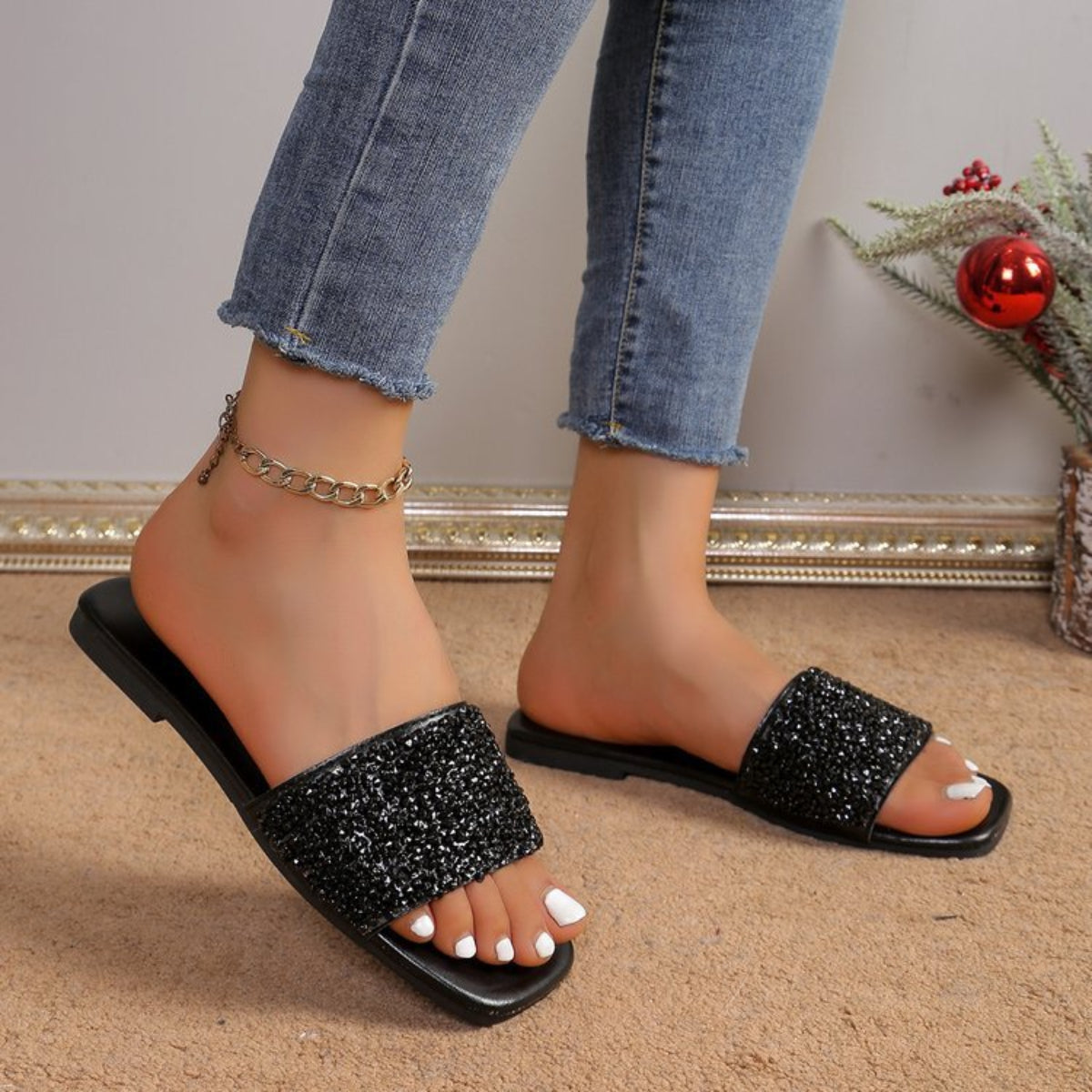 Black women’s rhinestone embellished flat slide sandals shown worn from an angled side view, featuring a wide glittering black upper strap, open-toe silhouette, matching black footbed, and slim flat sole styled with cropped denim for an elevated casual look.