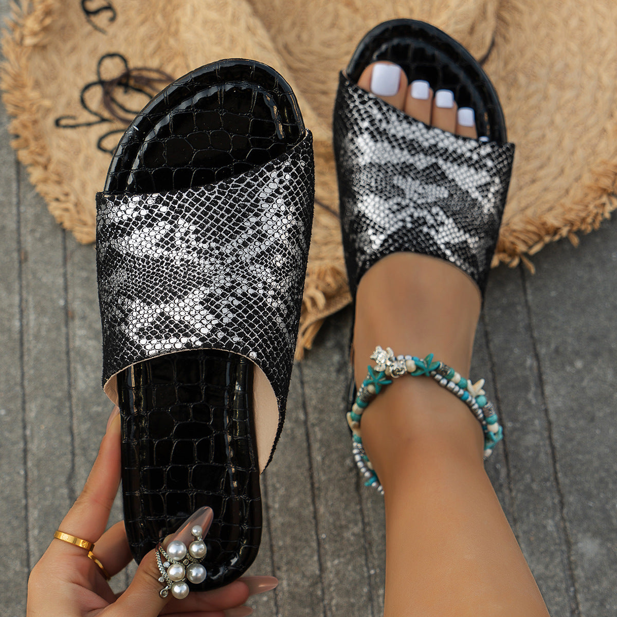 Black women’s flat slide sandals featuring a textured snake-print upper with subtle reflective detailing, open-toe construction, matching dark footbed, and low flat sole, shown from above with one sandal clearly displaying the patterned upper and overall slip-on shape.
