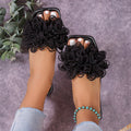 Black statement flat slide sandals for women featuring full layered 3D floral embellishments across the wide upper straps, open-toe styling, and a low flat sole, shown worn close-up with both feet visible to display the texture and volume of the flowers.