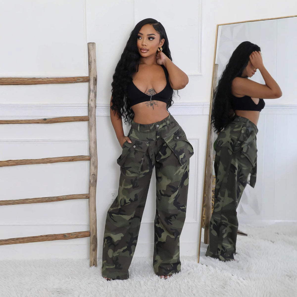Full-body front view of Briana women's high-waisted camouflage cargo pants modeled with a fitted black halter-style crop top, showcasing the oversized utility pockets, relaxed wide-leg fit, and floor-length hems. The pants feature an olive green, charcoal, and black military-inspired camouflage pattern, styled in a bright indoor setting with a wooden ladder and full-length mirror.