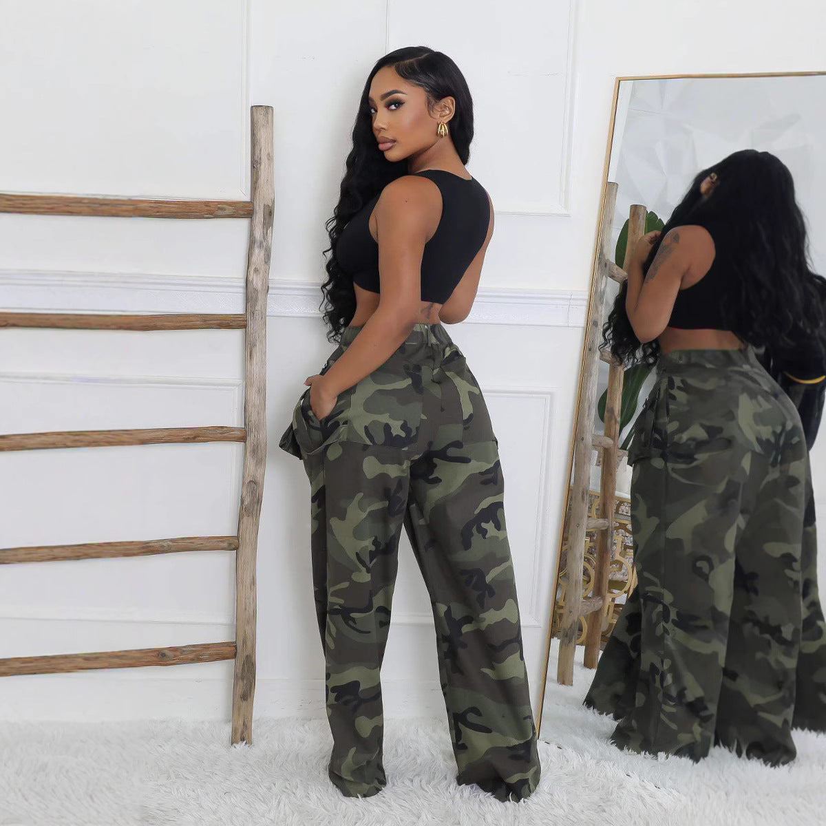 Three-quarter side view of Briana women's green camouflage cargo pants displayed on a model wearing a black halter crop top, featuring a fitted high waist, prominent oversized flap pockets at the hips and thighs, relaxed fabric through the legs, and wide floor-length hems. The model poses sideways beside a full-length mirror, showing the pants' utility-inspired construction and camouflage pattern.