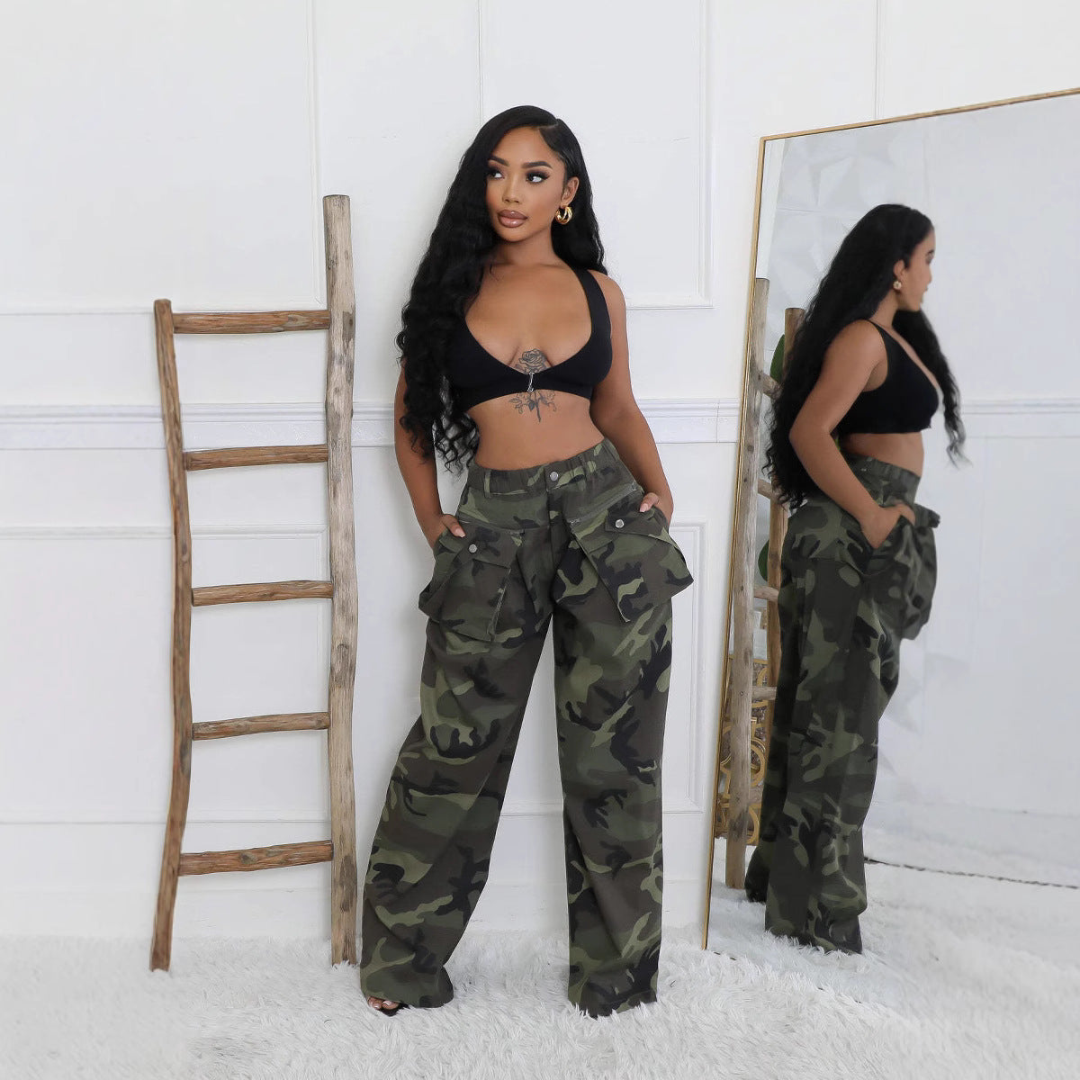 Briana women's green camouflage wide-leg cargo pants displayed in front and side views on a model wearing a black cropped halter top, featuring a high-waisted design, oversized flap cargo pockets along the thighs, a relaxed wide-leg silhouette, and an olive green, dark green, and black camouflage pattern. The model stands in a white room beside a wooden ladder and full-length mirror.