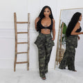 Briana women's camouflage wide-leg cargo pants shown from the front as a model steps forward wearing a black cropped top, highlighting the high-rise waistband, layered oversized thigh pockets, loose-fitting pant legs, and bold green and black camouflage print. The full-length pants drape over the model's footwear, creating a relaxed streetwear silhouette.