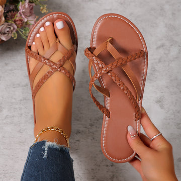Dark brown women’s braided cross-strap toe-ring flat sandals with woven straps crossing over the foot, open-toe styling, coordinating brown flat footbed, and contrast stitching around the sole, shown with one sandal worn and the matching sandal held for a clear product view
