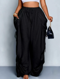 Delilah women's black high-waisted ruched wide-leg cargo pants shown from the front on a model wearing a white cropped top, featuring a gathered elastic waistband with adjustable drawstring ties, oversized draped side pockets, decorative ruching along the outer legs, and a relaxed, flowing full-length silhouette. The model holds a small black handbag with a gold chain strap against a light gray studio background.