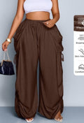 Delilah women's brown high-waisted wide-leg cargo pants displayed from the front on a model wearing a fitted white cropped top, featuring an adjustable drawstring waistband, oversized side cargo pockets, gathered fabric along the outer legs, adjustable ruched detailing near the lower hems, and a relaxed floor-length silhouette.
