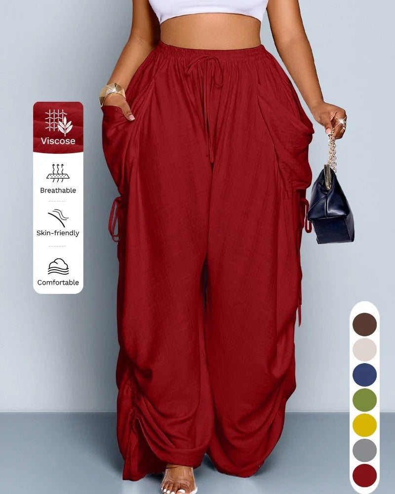 Delilah women's wine red wide-leg cargo pants shown full length on a model, highlighting the gathered high-rise elastic waistband, front drawstring tie, oversized utility-inspired pockets along both sides, draped fabric through the thighs, and long relaxed pant legs with decorative ruched detailing.