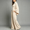 Side view of the Eliana women's apricot high-neck jumpsuit, highlighting the dramatic cape-style sleeves draping from the shoulders, fitted waistline, softly flowing fabric, and relaxed floor-length wide-leg pants.