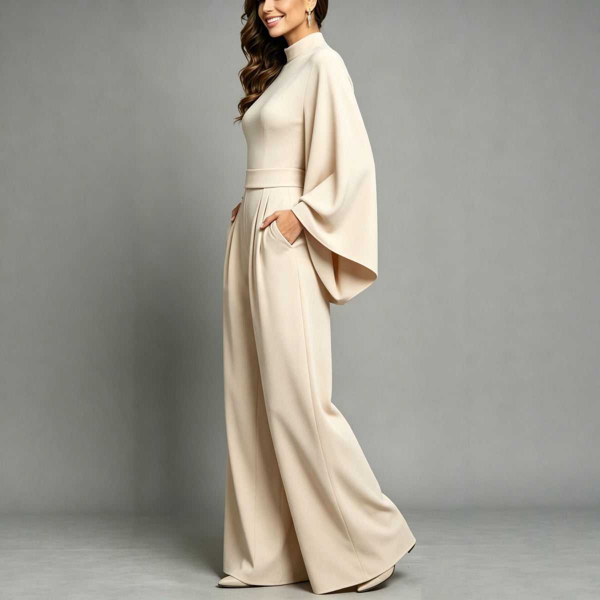 Side view of the Eliana women's apricot high-neck jumpsuit, highlighting the dramatic cape-style sleeves draping from the shoulders, fitted waistline, softly flowing fabric, and relaxed floor-length wide-leg pants.