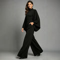 Eliana women's black high-neck jumpsuit shown on a model in a walking pose, featuring dramatic draped cape-style sleeves, a fitted waist, flowing wide-leg trousers, and an elegant full-length silhouette.