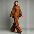 Eliana women's caramel elegant jumpsuit shown on a model in a walking pose, featuring a fitted high neckline, flowing cape-inspired long sleeves, a defined waist with front pleats, side pockets, and wide-leg trousers that drape toward the floor.
