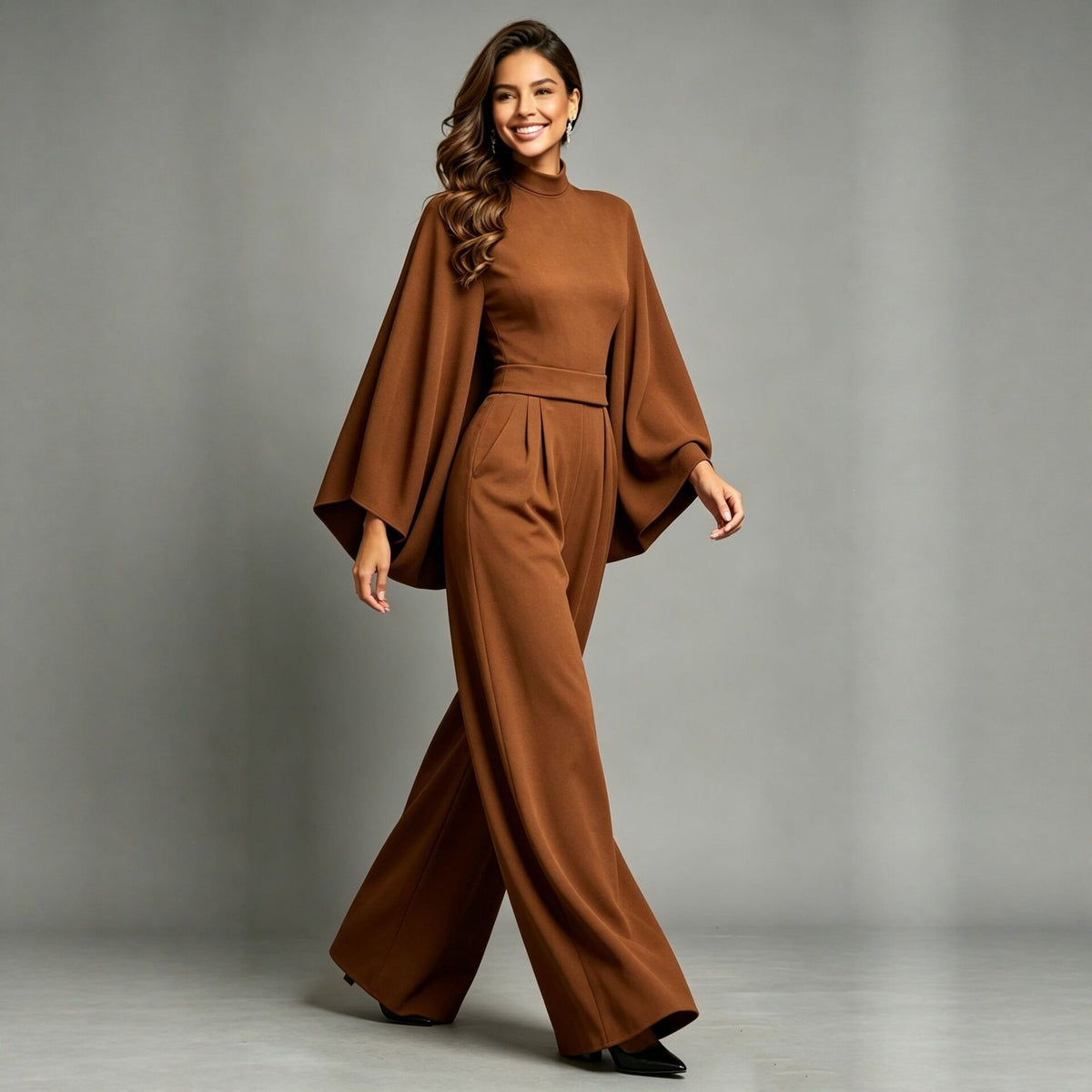 Eliana women's caramel elegant jumpsuit shown on a model in a walking pose, featuring a fitted high neckline, flowing cape-inspired long sleeves, a defined waist with front pleats, side pockets, and wide-leg trousers that drape toward the floor.