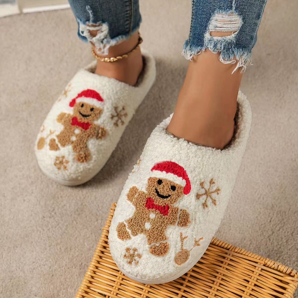 Women’s cream gingerbread Christmas house slippers shown worn while standing, featuring soft plush closed-toe uppers, embroidered gingerbread characters, festive red accents, brown snowflake designs, and cozy cream interiors for Christmas morning and winter lounging.