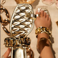 Gold women’s flat slide sandals featuring a metallic quilted upper, oversized polished gold-tone chain embellishment, open-toe silhouette, and flat sole, shown with one sandal held upright beside the matching sandal worn on the foot to highlight the metallic finish, textured upper, and statement chain detail.