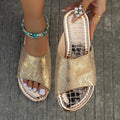 Gold women’s flat slide sandals featuring a shimmering snake-print textured upper, open-toe silhouette, low flat sole, and metallic gold finish, shown worn from a close top angle with white pedicure and turquoise ankle jewelry for a detailed view of the patterned upper and foot coverage.