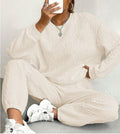 Harper women's cream cable-knit matching set displayed on a seated model, featuring a loose-fitting long-sleeve crewneck sweater with textured diamond-knit patterns, coordinating jogger pants with a relaxed tapered fit, and white chunky sneakers.