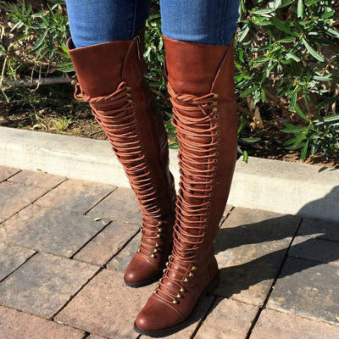 Woman wearing brown Harper cross-strap knee-high boots outdoors, featuring a tall fitted shaft, layered strap detailing, rounded toe, and comfortable block heel styled with blue jeans.