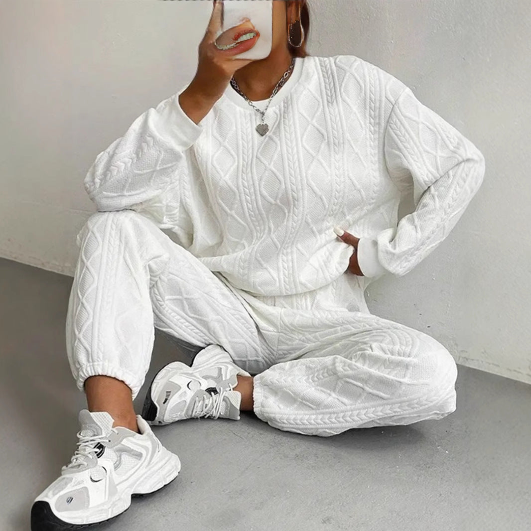 Harper women's white cable-knit two-piece lounge set shown on a seated model, featuring a relaxed long-sleeve crewneck sweater with raised diamond and braided knit patterns, matching drawstring jogger pants with textured detailing, and chunky white athletic sneakers.