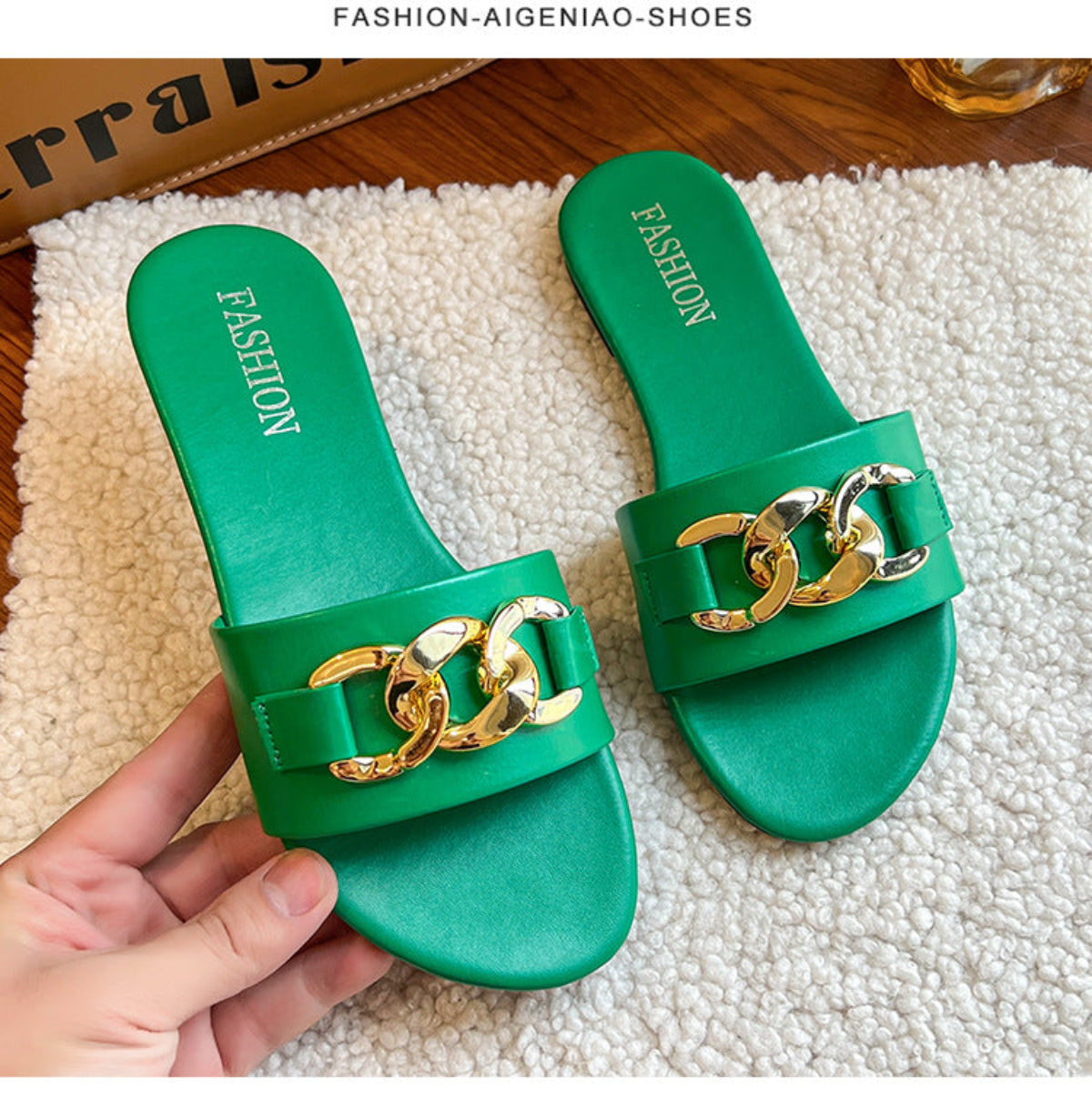 Kelly green women's flat slide sandals with a bold green strap, oversized polished gold-tone chain buckle embellishment, open-toe design, and matching green footbed, displayed as a pair on a soft cream textured rug.