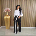 Kendra women's black-and-white high-waisted wide-leg pants shown from the front on a model wearing a white cropped button-up blouse, featuring a striking asymmetrical white stripe beginning at the waistband and extending vertically down one leg, a fitted-looking waist, and relaxed full-length wide-leg trousers. The model stands beside a tall gold decorative vase filled with pale pink flowers against a vertical wood-slat wall.