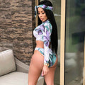 Side and three-quarter back view of a woman modeling a lavender tropical, mint green, and white floral-print two-piece swimsuit. The swimwear features a fitted long-sleeve cropped top with a rounded neckline, coordinated high-cut bikini bottoms, and a matching printed headband. The tropical-inspired floral design combines lavender purple flowers, mint green leaves, and a soft white background, creating a colorful beachwear look.