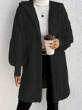 Woman wearing a black hooded teddy longline coat over a white turtleneck and black pants while holding a coffee cup against a light neutral wall.