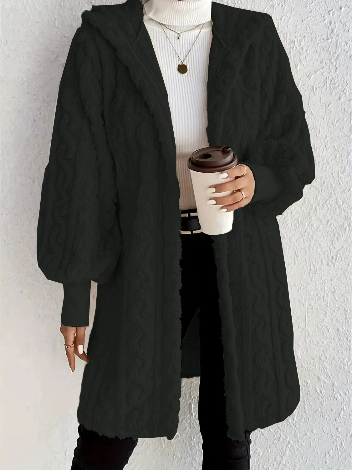Woman wearing a black hooded teddy longline coat over a white turtleneck and black pants while holding a coffee cup against a light neutral wall.