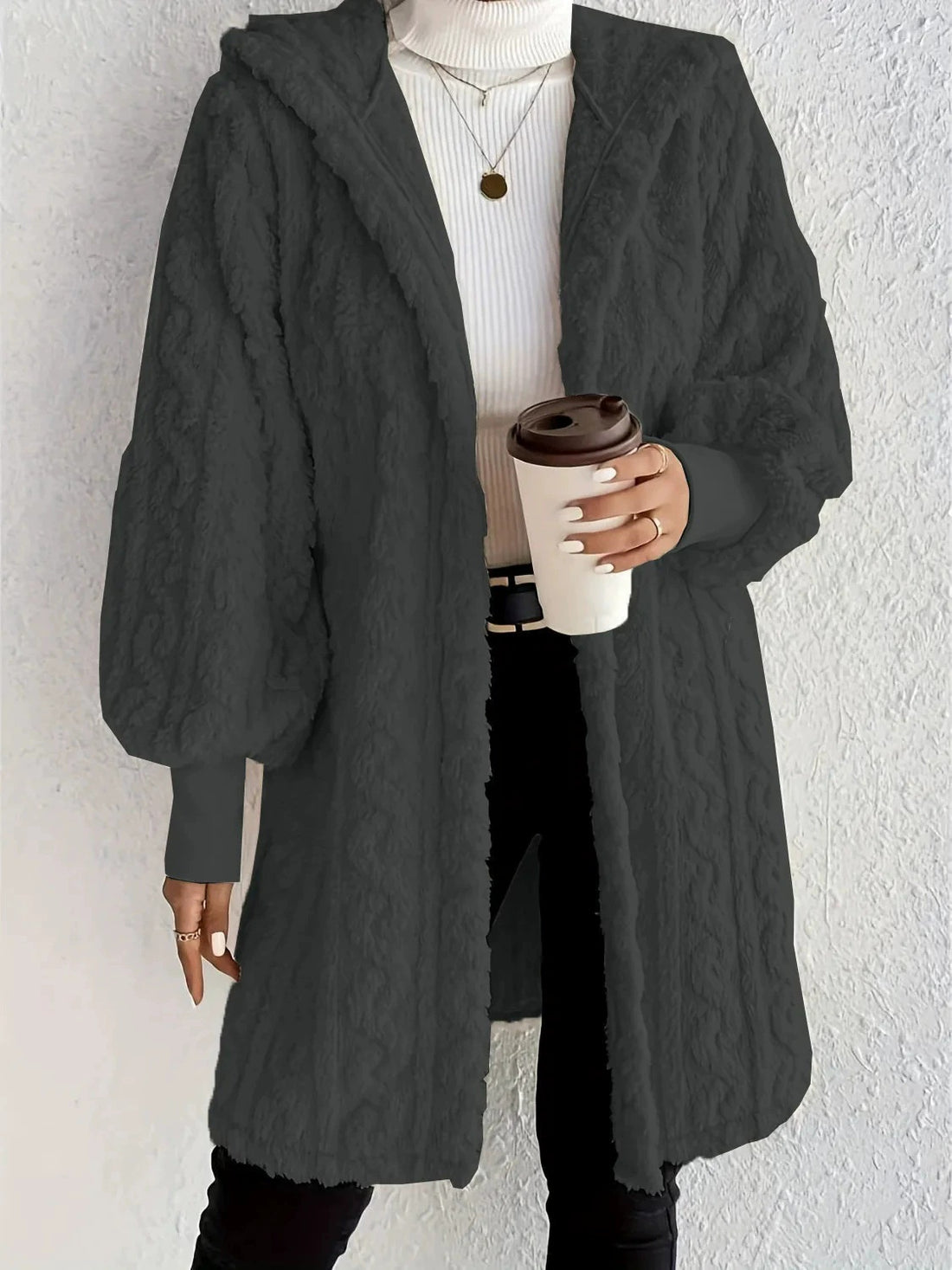 Woman wearing an white milk hooded teddy longline coat over a white turtleneck and black pants while holding a coffee cup against a light neutral wall.