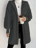 Woman wearing a dark grey hooded teddy longline coat over a white turtleneck and black pants while holding a coffee cup against a light neutral wall.