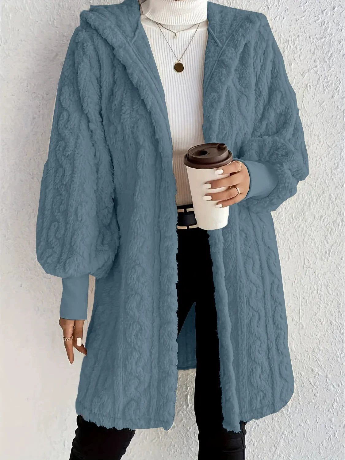 Woman wearing an ocean blue hooded teddy longline coat over a white turtleneck and black pants while holding a coffee cup against a light neutral wall.