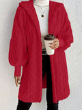 Woman wearing a red hooded teddy longline coat over a white turtleneck and black pants while holding a coffee cup against a light neutral wall.