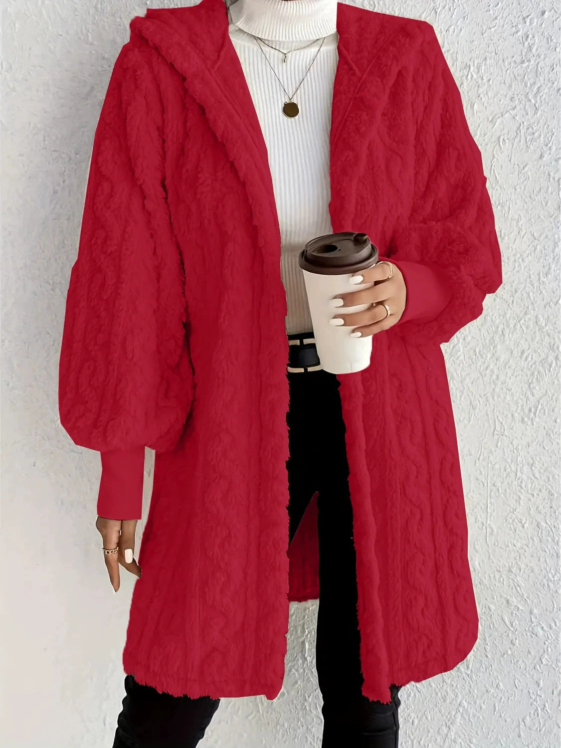 Woman wearing a red hooded teddy longline coat over a white turtleneck and black pants while holding a coffee cup against a light neutral wall.