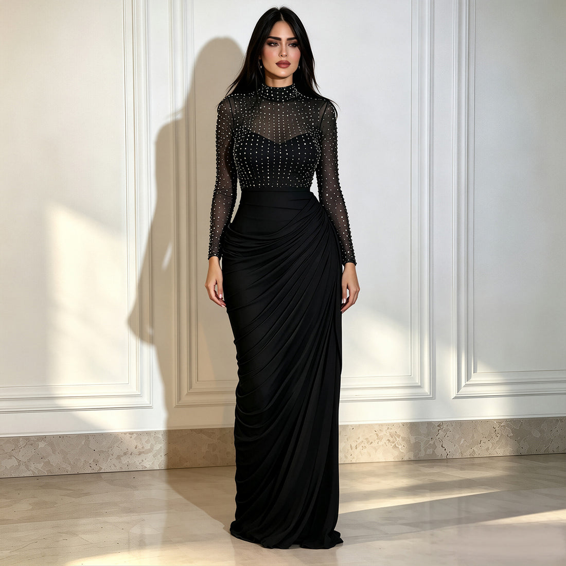 A woman modeling a long black evening gown with a sheer embellished mesh bodice and a draped floor-length skirt, standing against a white paneled wall.