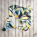 Women's two-piece swimwear set displayed flat on a rustic wooden background, featuring a long-sleeve cropped swim top and matching bikini bottoms. The swimsuit showcases a bold abstract geometric pattern with sweeping curves and stripes in turquoise blue, yellow, white, black, and deep teal. The cropped top features a fitted silhouette and long sleeves, while the coordinating bikini bottoms feature matching multicolor patterns.