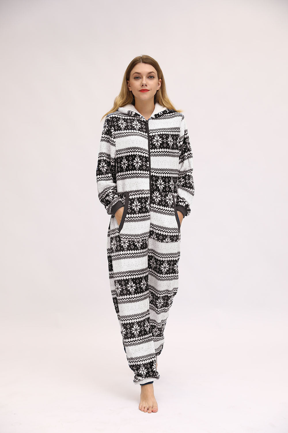 Full-length front view of the Noelle black and white hooded pajama onesie worn by a woman standing against a light studio background, featuring black reindeer silhouettes, white snowflake and winter-inspired geometric patterns, a front zipper, attached hood with drawstrings, and tapered full-length legs with gathered cuffs.