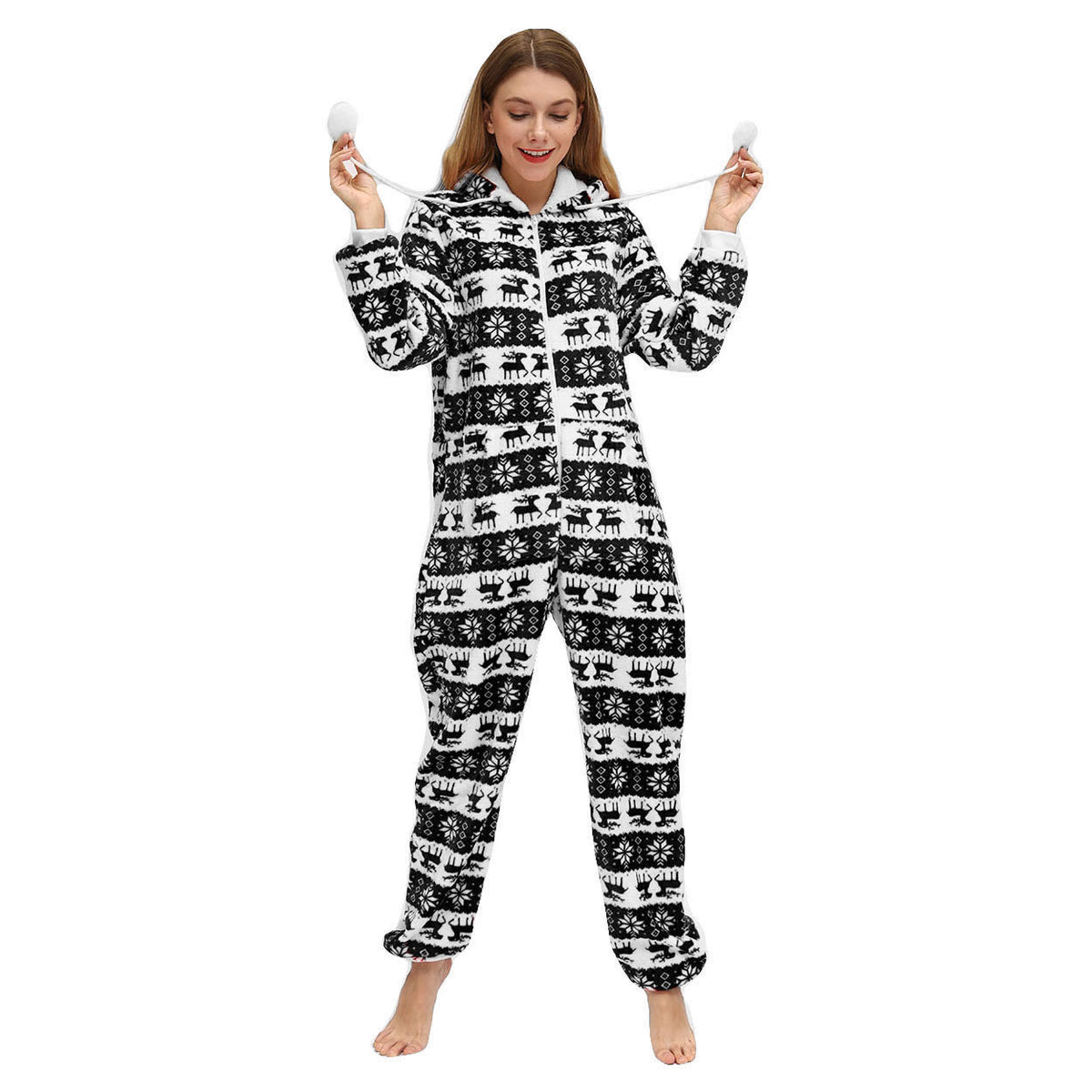Full-length angled view of a woman wearing the Noelle black and white Christmas pajama onesie, featuring horizontal rows of black reindeer, snowflake designs, geometric winter patterns, a zippered front, fitted-looking hood, long sleeves with elasticized cuffs, and matching full-length pajama legs gathered at the ankles.